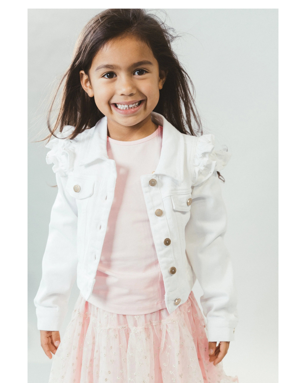 Young girl wearing a white jean  jacket over a pink dress on a light gray background